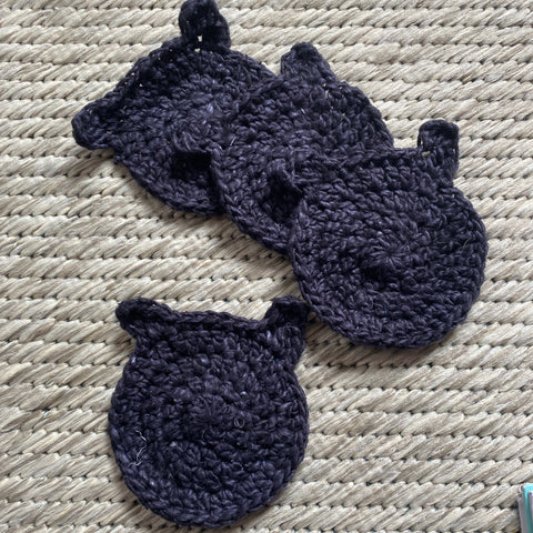 Against a warm tan rug, four different coasters made out of black yarn with little cat ears are posed.