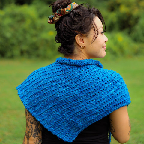 A woman with long black hair in a bun is facing away from the camera, wearing a bright blue knitted shawl over her shoulders.