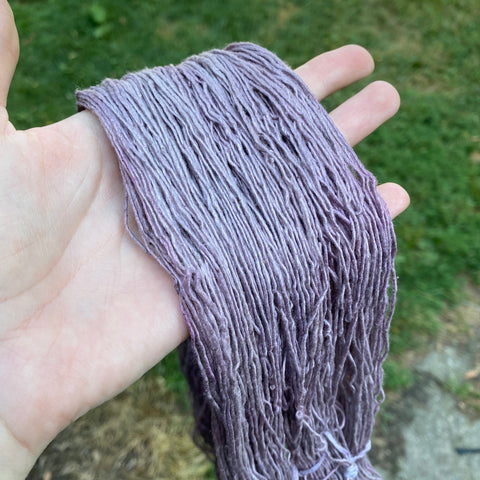 A hand holding out an unwound skein of dyed yarn. The white yarn is now a few shades of greyish purple.