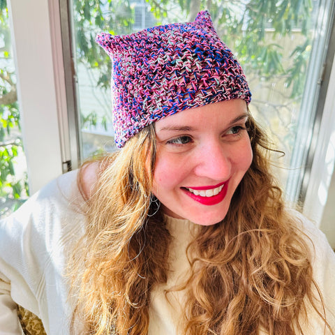 Woman wearing red lipstick looking away from camera smiling and wearing purple cat ear beanie in front of window full of sunlight.