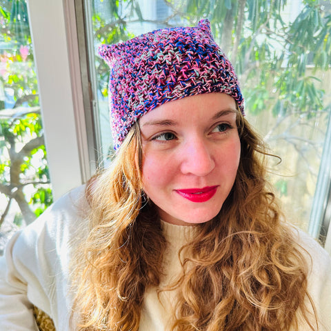 Woman wearing red lipstick looking away from camera wearing purple cat ear beanie in front of window full of sunlight.