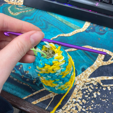 The teal and yellow body of the fish is hanging off a purple crochet hook and being stuffed with a mix of polyfill and catnip.