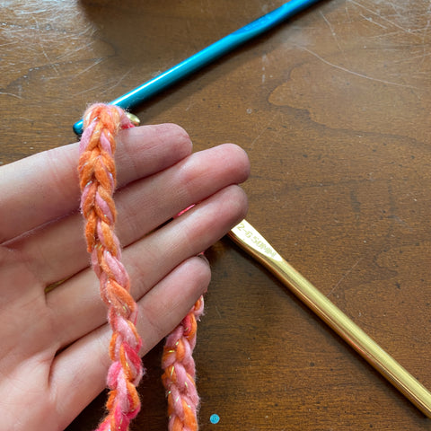 On a wooden table, a blue and a gold metal crochet hook rests on the table. In a pale hand is a chunky crochet chain of pink and orange ombre sparkle yarn.