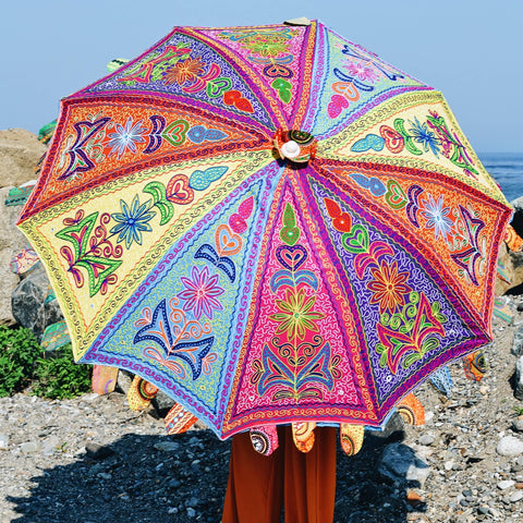 A person is covering themselves up with a multicolored, brightly colored embellished and embroidered umbrella