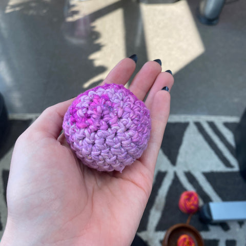 A hand outstretched, holding a small amigurumi circle made of sparkly ombre pink yarn.