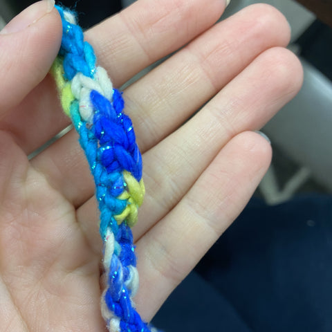 A hand is holding a crochet chain, made out of blue, teal, white, yellow, and sparkly yarn.