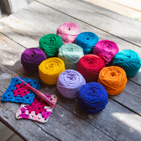 On a gray picnic table, 12 skeins of brightly colored yarn in all different colors are arranged neatly into a square. To the left of the yarn are two granny squares made out of blue and pink yarn, and a pink crochet hook.
