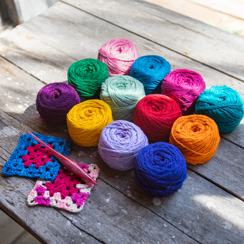 On an aged picnic table lay 12 different colored skeins of solid colored worsted weight silk yarn. There is also a pink crochet hook and two granny squares, made of different blue and pink yarn.