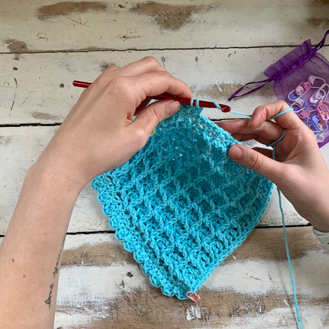 A pair of hands are working on a washcloth made out of teal yarn, using the waffle stitch