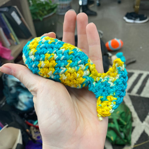 A hand is outstreched, holding an amigurumi fish made of teal, white, and yellow worsted weight silk yarn.