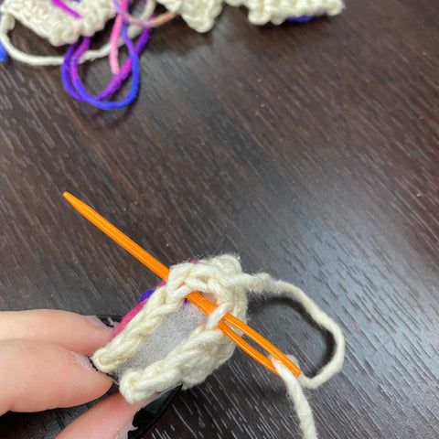 A  hand is holding onto the two sides of a rune and is sewing it together with an orange yarn darning needle.