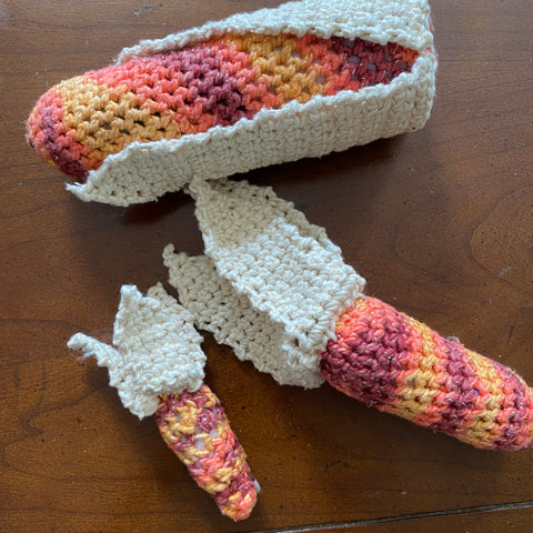 A large ear of crochet fall-colored corn is wrapped in it's white husk on a wooden table. Beneath the large cob are two smaller cobs, their white husks pulled back away from the cob.