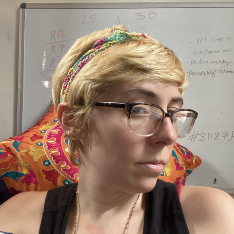 A person with short blonde hair and a black tank top and glasses is posing in front of a white board and a decorative orange and pink pillow. On their head is a crochet headband made out of sparkling lace weight silk yarn.