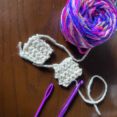 Two blocks made of white yarn are laying on a dark wooden table, next to a purple crochet hook, a purple darning needle, and a skein of pink-blue-white multicolored worsted weight silk yarn.