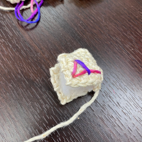 A rune made of white and multicolored yarn is half sewn, open and showing the stuffing inside.