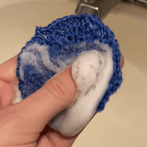 A gif of a thumb swirling white foamy suds of soap around the circle of the blue reusable crochet make up remover pad.