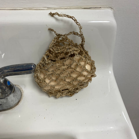 A bar of soap that is held securely in a handmade crochet hemp soap saver on the edge of a white sink.