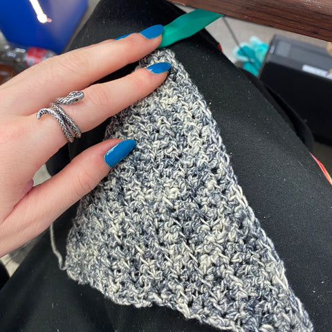 A hand with blue nails is holding onto a small triangle made from white and grey speckled yarn.