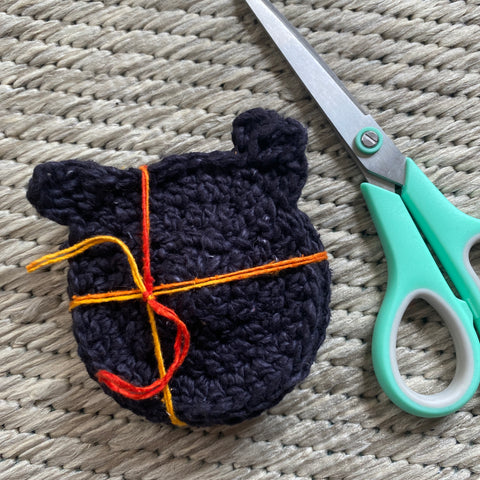 Against a tan rug, black cat coasters are knotted together with a strand of orange thread. To the right of the coasters is a pair of green and silver scissors.