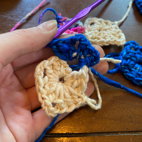A pale hand is holding a half finished granny square, the first corner of the square filled in with sparkle classic blue worsted weight yarn.