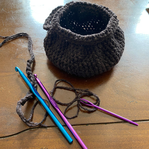 On a shiny wooden table, a black crochet witch's cauldron sits finished beside a few strands of black yarn, a blue and purple crochet hook, and a purple darning needle.