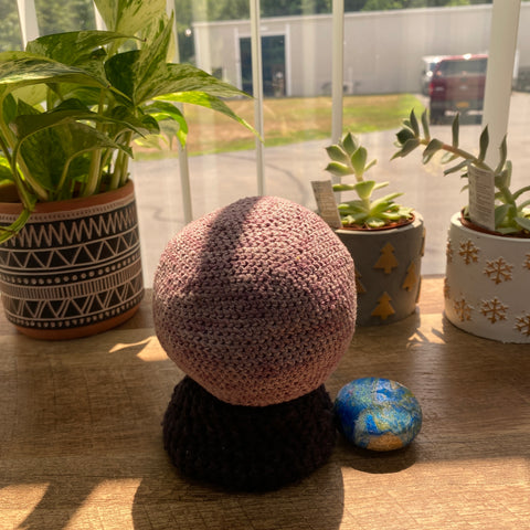 A crystal ball made of light smoky purple and black yarn is resting on a wooden table in the sun. Behind the ball are a few different types of green, lush plants.