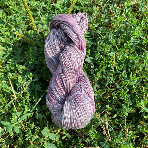 A purple-ish gray skein of yarn is resting in the grass in the sunlight, showing off the different shades of purple and soft lavender.