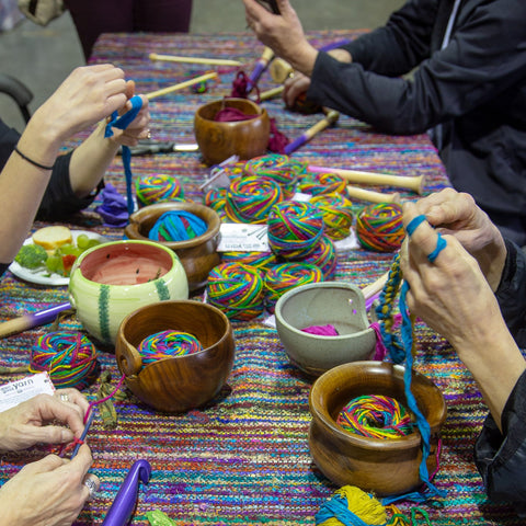 A table with a rainbow table cloth is covered in yarn bowls, knitting needles, crochet hooks, and rainbow yarn. People are sitting around the table, their hands hard at work knitting and crocheting.