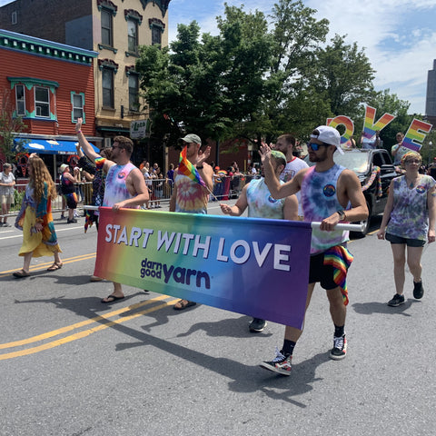 The whole DGY crew, wearing tie dye shirts and holding a rainbow 'START WITH LOVE - DARN GOOD YARN' banner, are waving as they march down the street at the Albany Pride Parade 2022
