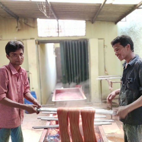 Two men are holding metal dyeing poles. On the middle of the poll are some hanks of yarn that are being dipped into steaming hot liquid herbal dye.