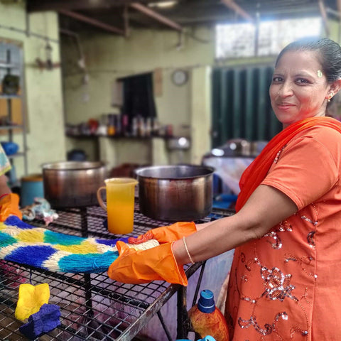 An Indian woman wearing an orange sparkly sari is wearign a pair of orange gloves as she dyes white silk yarn with blue, teal, and yellow dyes.