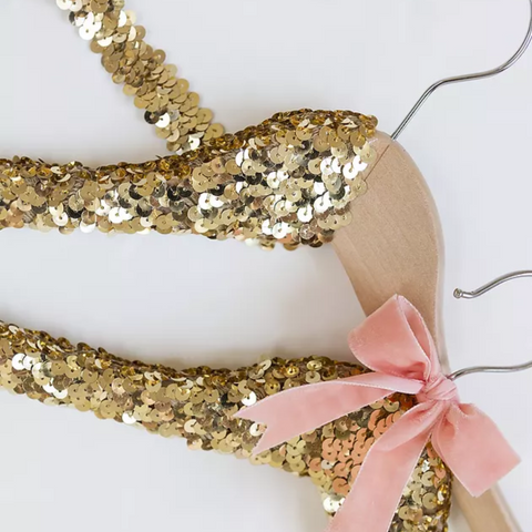A pair of wooden hangers are being wrapped in golden sequins. A pink satin bow is wrapped around the hook of one of the hangars. 