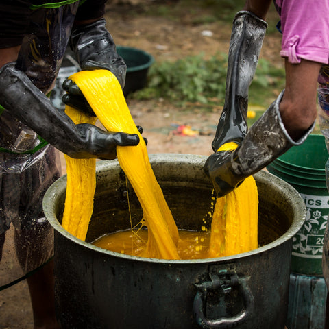 Two people wearing thick black gloves and aprons are pulling out large hanks of bright yellow yarn out of a warm dyeing pot.
