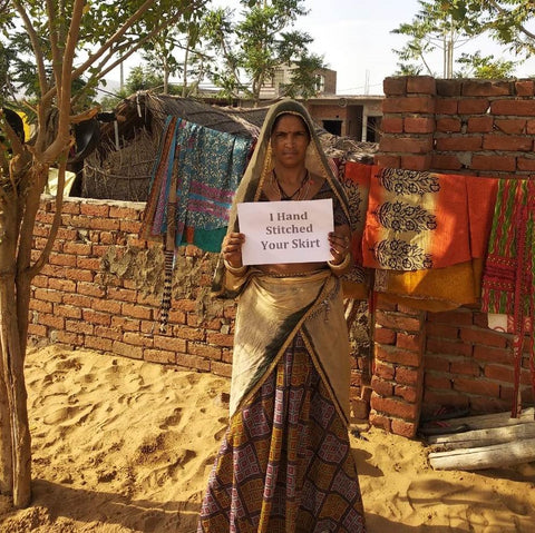 A woman wearing a tan and red sari is standing outside by a brick wall. Behind her is a clothes line where sari wrap skirts hang. In the woman's hand is a white sign reading 'I hand stitched your skirt'