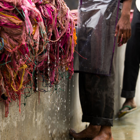 A man wearing a clear apron and dark pants is standing barefoot by a large cement wall. On the ledge of the wall is a huge pile of dripping pink yarn. This is yarn that has just been rinsed and needs to be hung out to dry.