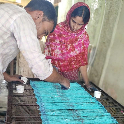 A man in a white shirt nd a woman wearing a pink sari are sprinking purple dye powder onto teal yarn that is streched over a metal rack.