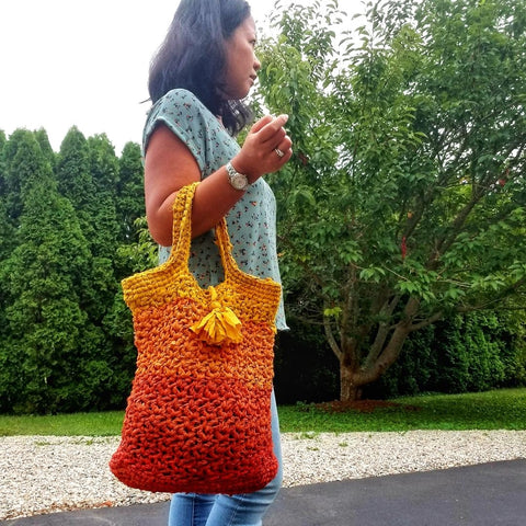 A hispanic woman with dark hair, blue jeans, and a blue shirt. She's walking in the woods and holding an orange ombre crochet tote in her elbow. 