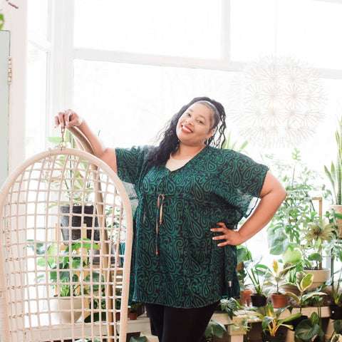 A plus sized woman of color wearing black leggings and a green tunic is leaning against a white wicker seat in a brightly lit green house