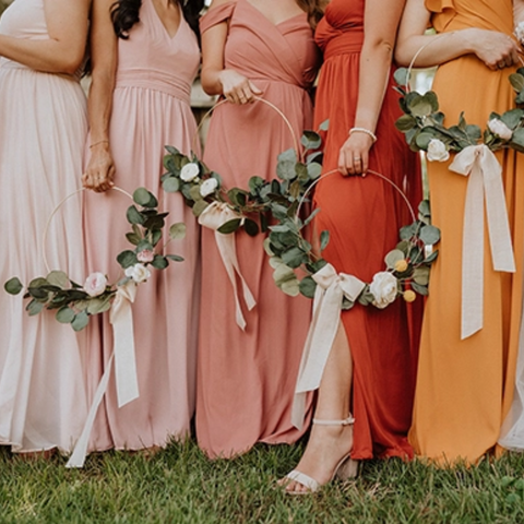 5 women, all wearing different warm shades of pink/orange, are posing outside, each holding a golden hoop adored with flowers, leaves, and white ribbons. 