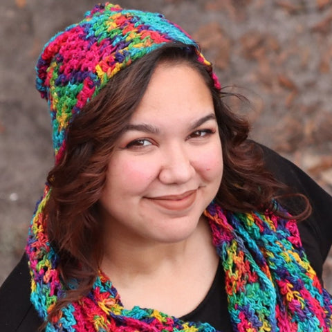 A plus sized woman with wavy brown hair is smiling up at the camera, wearing a hat and scarf made of rainbow worsted weight watercolors silk yarn.