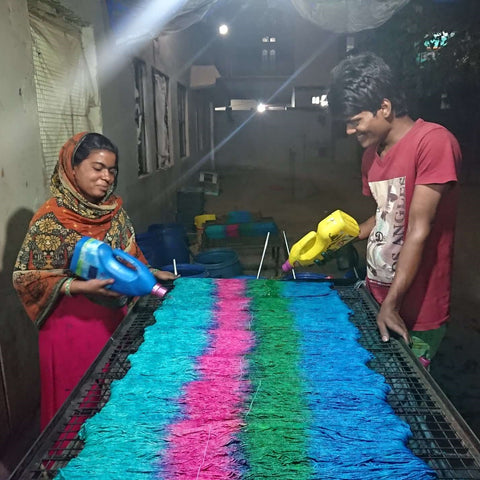 Two Indian artisans are standing at opposite ends of the yarn dyeing table. Each artisan has a bottle of dye in their hands that they are using to dye the yarn blue, green, and pink.