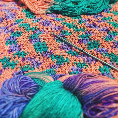 A close up of a piece of crochet work, made up of pink, purple, and teal sport weight yarn. At the forefront of the picture is an unused skein of the same pink-purple-and teal sport weight yarn.