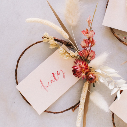 A circular name card, that says Katy in red writing is tied together with ribbon and dried flowers.