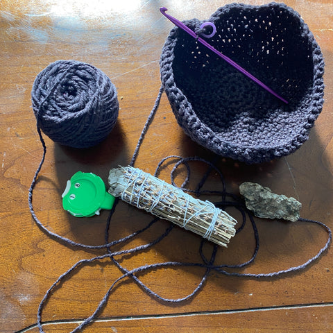 A cake of black yarn is still connected to the purple crochet hook, which is resting inside the half finished cauldron. Beside the work is a green stitch marker, a shiny rock, and a bundle of lavender sage.