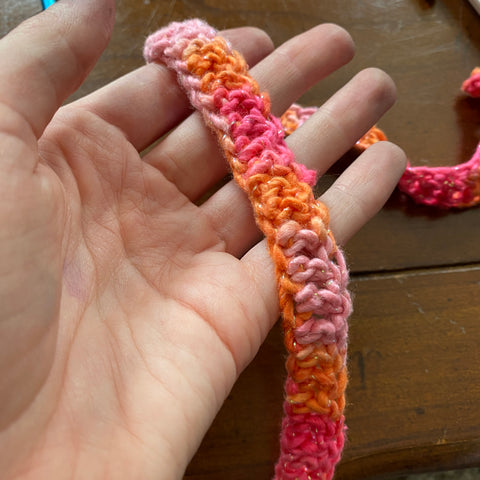 On a wooden table, a pale hand holds a strand of double crochet pink ombre sparkle worsted weight yarn.