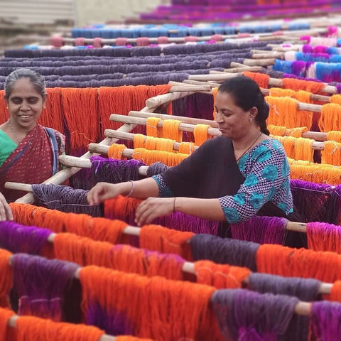 Two Indian women are standing outside, adjusting yarn on large wooden sticks so that they yarn can dry in the sun. There are dozens of these sticks, filled with loose skeins of orange, blue, purple, and pink yan.