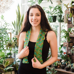 A girl with long dark hair wearing a black tank top and a colorful knit green scarf is smiling in a greenhouse, surrounded by plants.
