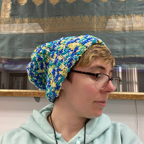 A person with glasses and short blond hair is wearing the blue, white, and yellow worsted weight beanie.