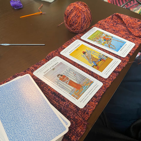 On a wooden table, a tarot mat made of orange and black yarn is spread out. A tarot deck and three tarot cards are spread out, ready to be read.
