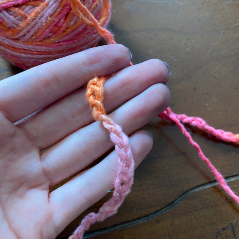 A pale hand is holding a chain of stitched rose quartz pink yarn. To the upper left of the hand is the full cake of the same pink sparkly yarm.